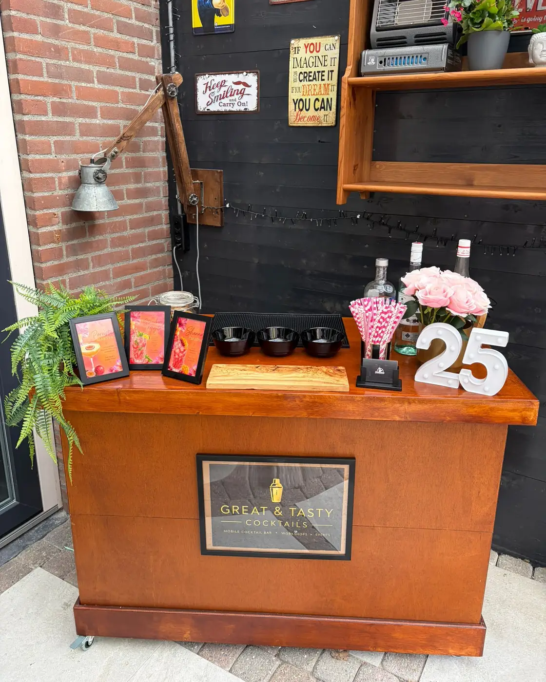 De mobiele cocktailbar frontaal, met drie ingelijste cocktailkaarten en garneringsbakjes op het werkblad en de wielen zichtbaar onder de plint
